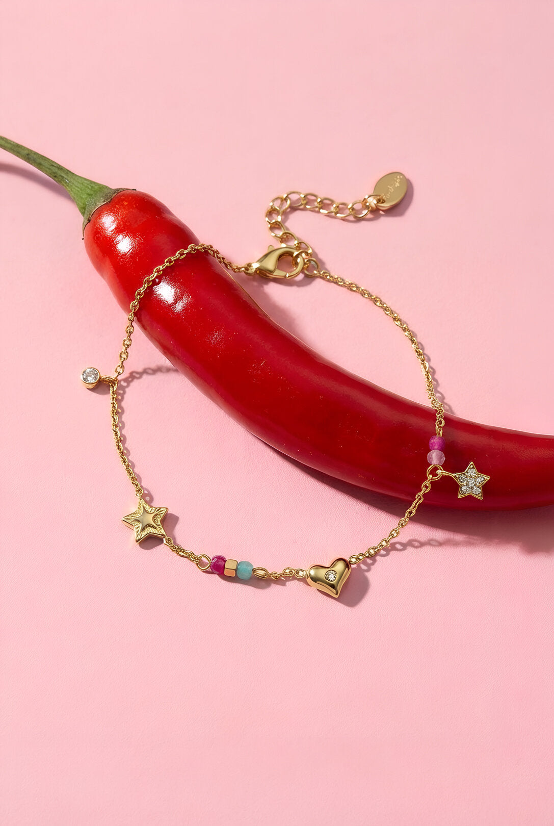 Braccialetto con ciondoli cuore stella pietre su sfondo rosa con peperoncino rosso.