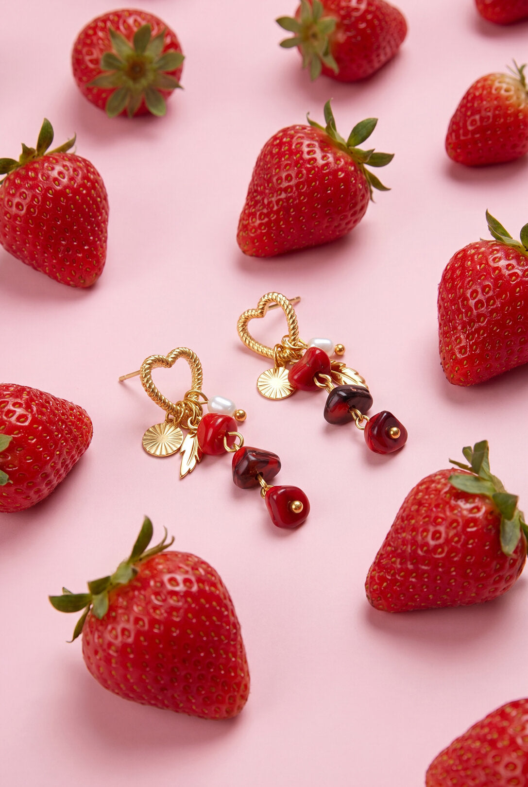 Orecchini cuore perla piuma con pendenti, circondati da fragole fresche su sfondo rosa.